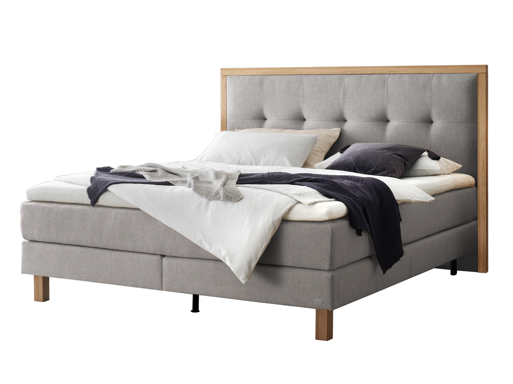 Ein freigestelltes Produktfoto des Boxspringbettes Hasena Pronto Solara mit hellgrauem Stoffbezug und Holzrahmen, dekoriert mit Decken und Kissen.