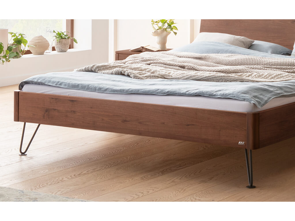 Nahaufnahme des unteren Teils eines Hasena Bett Locarno mit einem Rahmen in Walnuss-Holz (Nussbaum) und schwarzen, anthrazitfarbenen Haarnadel-Beinen. Man sieht den Bettrahmen, die Beine und den Holzboden.