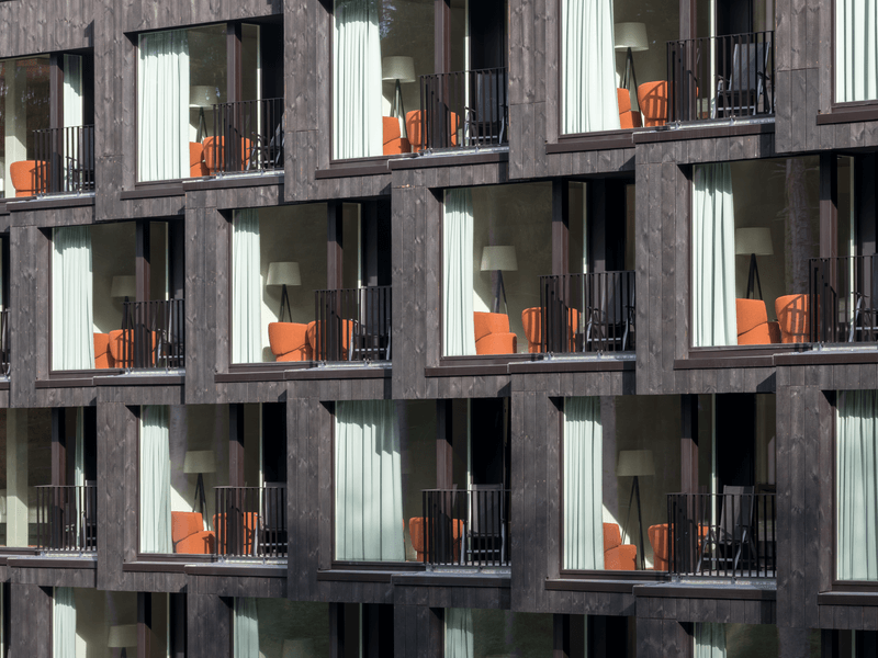 Nahaufnahme einer modernen Fassade mit mehreren Balkonen, auf denen orangefarbene Stühle und Lampen durch große Fenster sichtbar sind.
