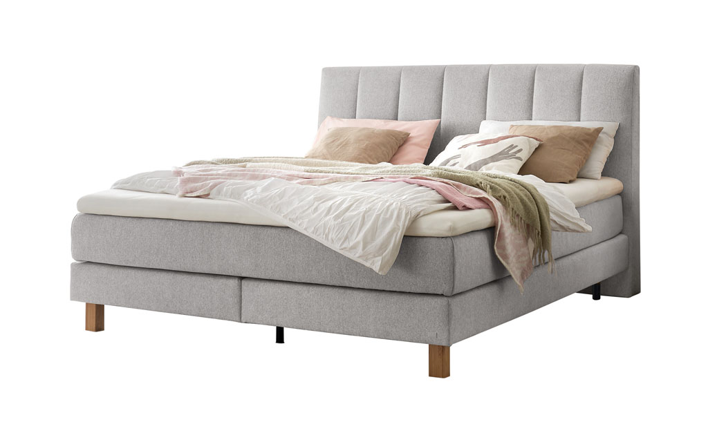 Ein Boxspringbett in einem hellen Grauton mit einem vertikal gesteppten Kopfteil, freigestellt auf weißem Hintergrund.