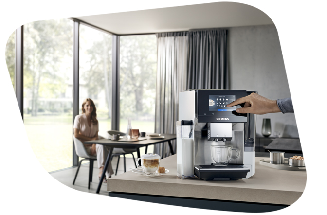 Ein Mann bedient einen modernen Siemens EQ Kaffeevollautomaten per Touchscreen in einer hellen Küche. Im Hintergrund sitzt eine Frau an einem Tisch. Eine Tasse Latte Macchiato steht auf der Ablage.