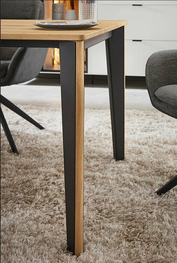 Detailansicht eines modernen Esstisches mit Holzplatte und schwarzen Metallbeinen auf einem beige Teppich, umgeben von grauen Stühlen.