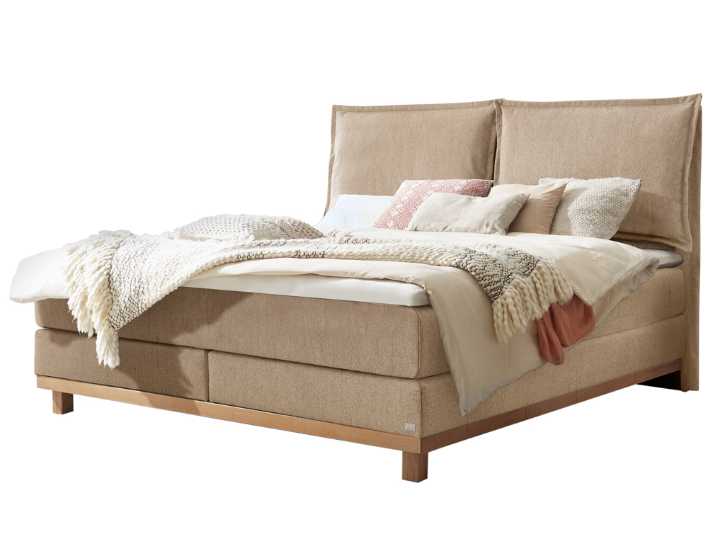 Freigestelltes, sandfarbenes Boxspringbett mit hellem Holzbettgestell und hoher, zweiteiliger Lehne.