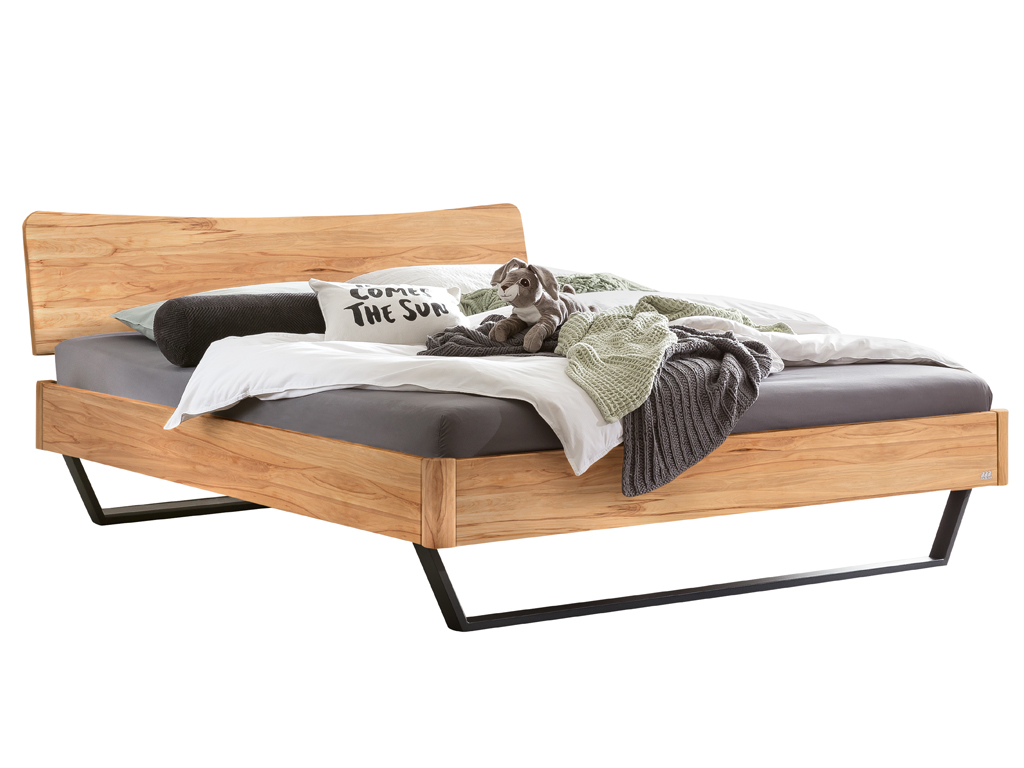 Ein Hasena Bettmodell Tresali aus Wildbuche mit ausgeprägter Maserung, einem geschwungenen Kopfteil und schwarzen Metallfüßen, auf dem Kissen und eine Decke liegen.