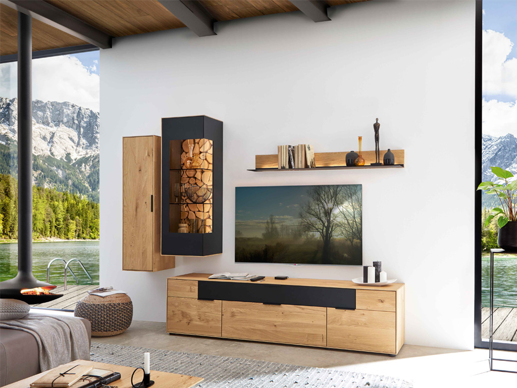 Wöstmann WM 2140 - Wohnwand  | Type 0002/0102 | Wildeiche massiv | konfigurierbar Ein stilvoll eingerichtetes Wohnzimmer mit Holzmöbeln und Dekoration, einem großen Fernseher, im Hintergrund ein beeindruckender Ausblick auf Berge und einen See.