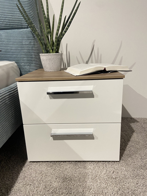 Rauch - Aldono | 2x Nachtkonsolen Eiche Weißer Nachttisch mit zwei Schubladen, dekoriert mit einem grünen Topfpflanze und einem geöffneten Buch, neben einem blauen Bett auf Teppichboden