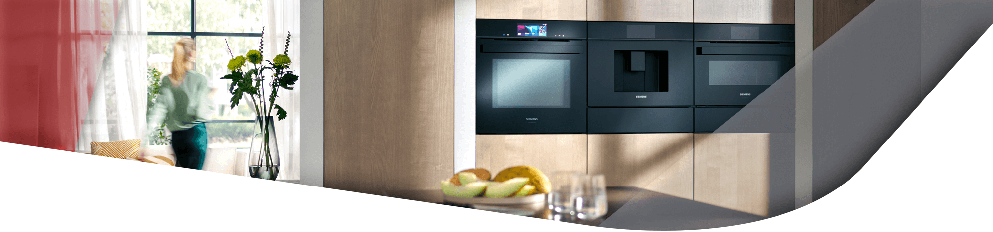 Einbauküche mit modernen Siemens Elektrogeräten, darunter ein Ofen und ein Kaffeevollautomat, daneben ein Tisch mit Obst und ein Fenster im Hintergrund.