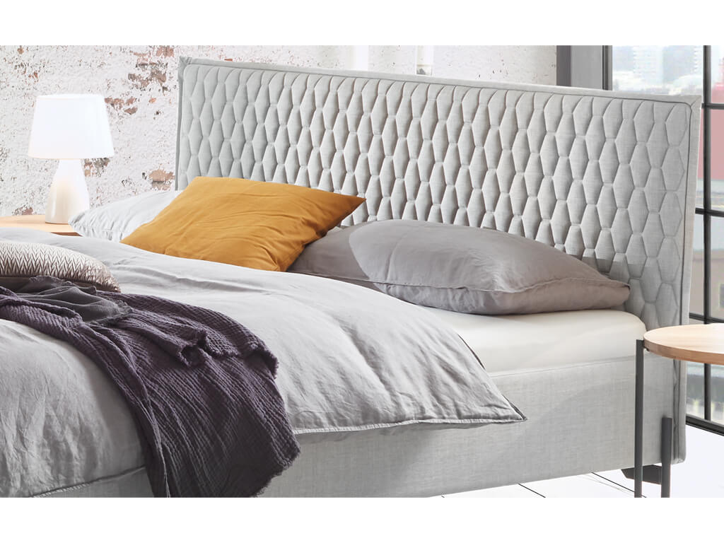 Eine Nahaufnahme des detailliert gesteppten, grauen Stoffkopfteils des Bettes Gloria, das mit Kissen und einer Decke für einen gemütlichen Look dekoriert ist.