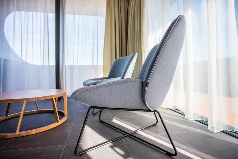 Ein moderner grauer Sessel mit schlankem Metallrahmen neben einem runden Holztisch vor bodentiefen Fenstern mit hellen Vorhängen