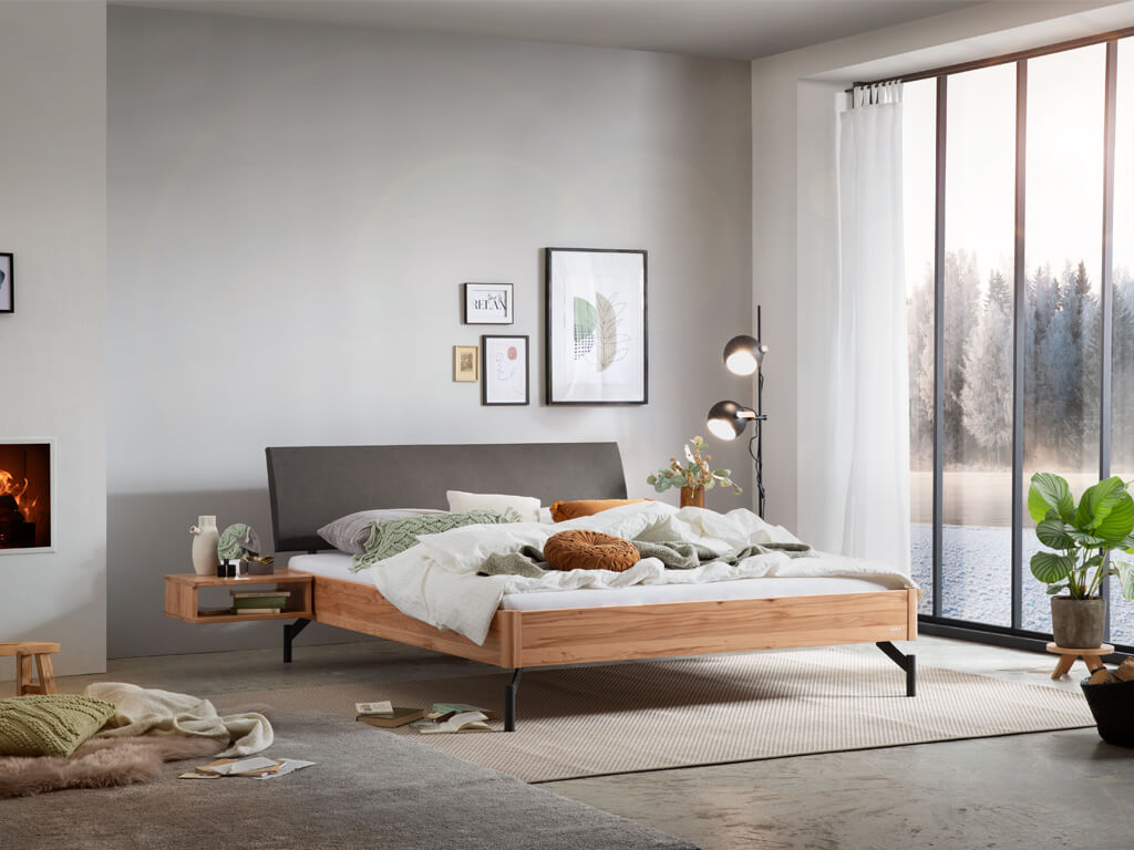 Eine Gesamtaufnahme eines Schlafzimmers mit dem Bettmodell "Broni". Das Bett hat einen Rahmen aus Wildbuchenholz, ein anthrazitfarbenes Kopfteil und schwarze Metallbeine. Ein Nachttisch aus dem gleichen Holz ist am Bett befestigt.