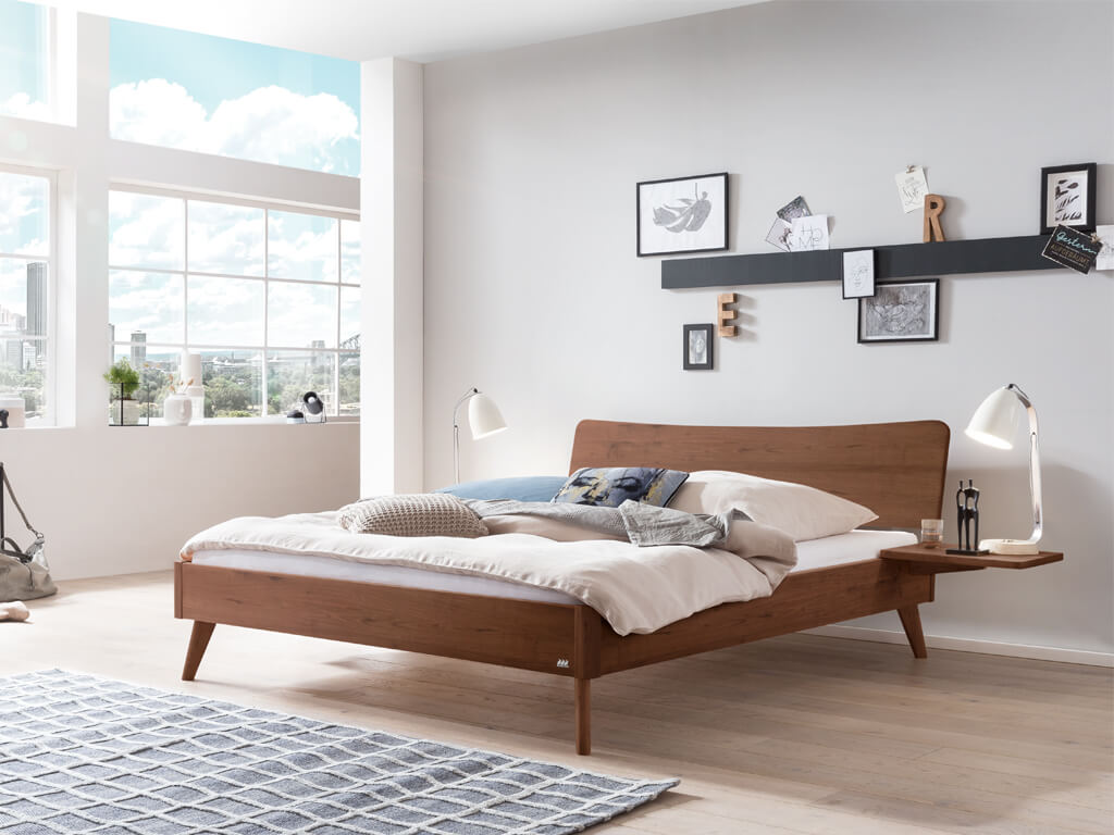 Ein Hasena Bett Caluso aus Nussbaum in einem hellen, modern eingerichteten Schlafzimmer mit großen Fenstern. Das Bett hat ein geschwungenes Kopfteil und schräg ausgestellte Beine.