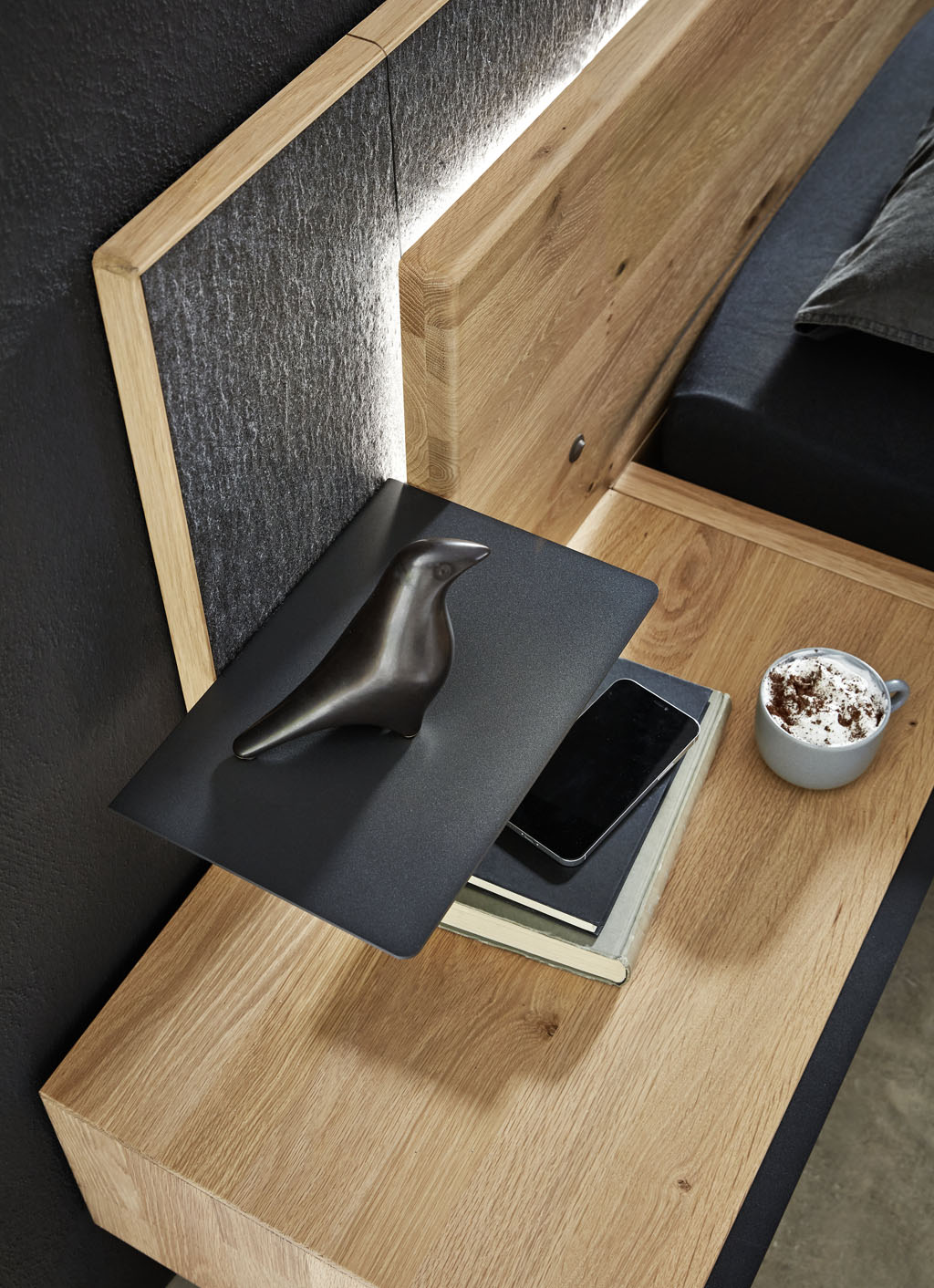 Detailaufnahme eines Bettes mit beleuchtetem Holzkopfteil und schwarzer Metallablage sowie einer grauer Kopfteilumrandung stilvoll dekoriert