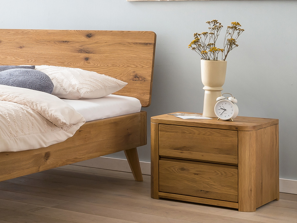 Detailaufnahme eines braunen Holzbettes mit weißer Bettwäsche einem Nachtschrank mit zwei Schubladen mit einer Vase und Wecker