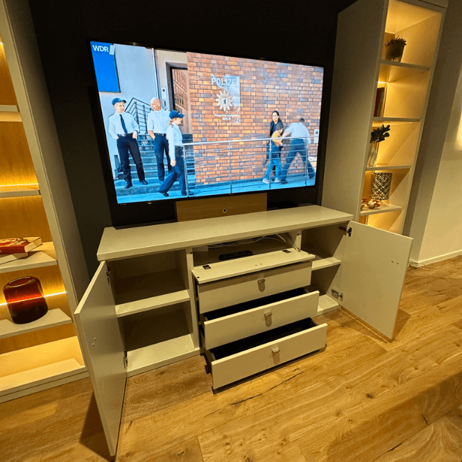 Wohnzimmerschrank mit offenen Schubladen und einem eingeschalteten Fernseher, der eine TV-Sendung zeigt, auf Holzboden und umgeben von Regalen