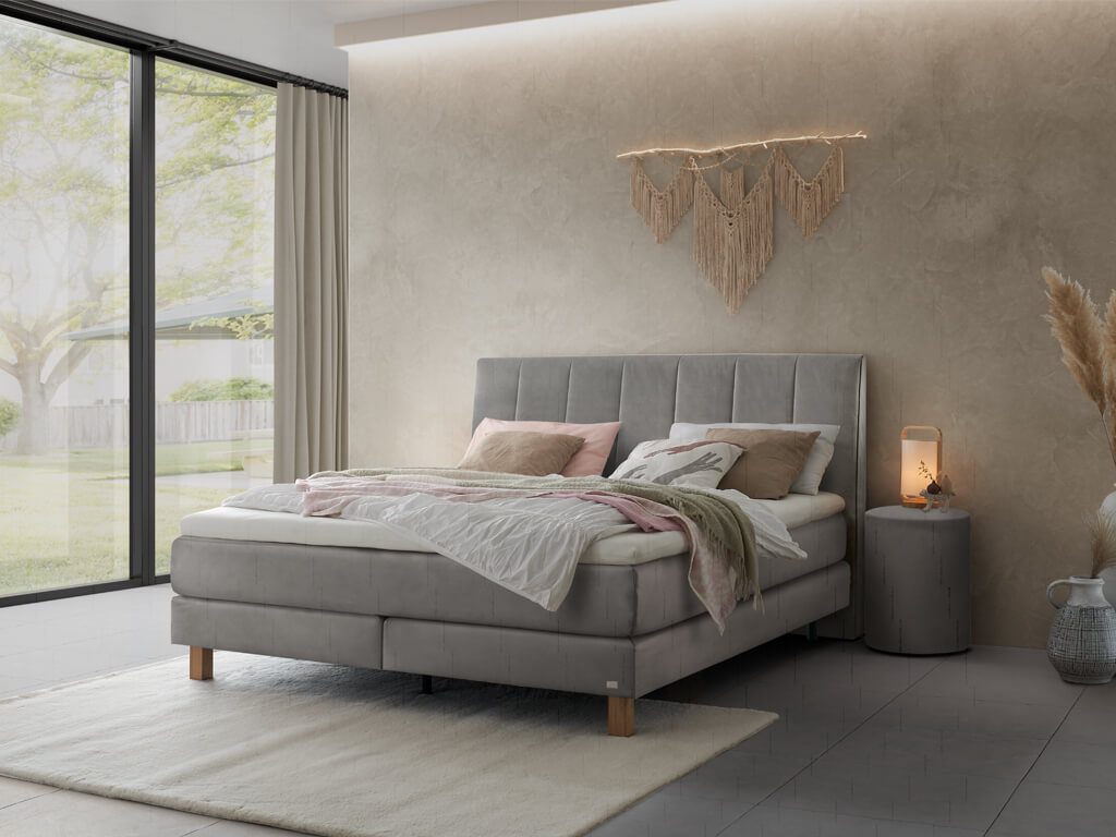 Ein modern eingerichtetes Schlafzimmer mit einem grauen Boxspringbett mit vertikal gestepptem Kopfteil in einer hellen Umgebung mit großer Fensterfront.