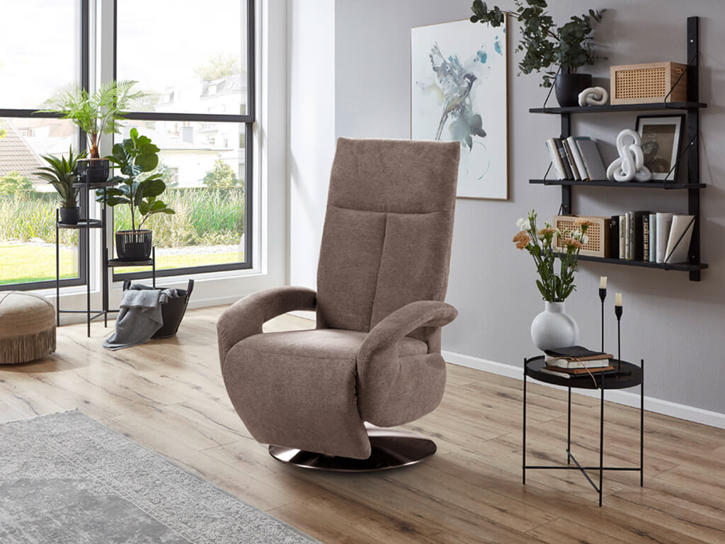 sit&more TV Tycoon 1222 | TV Sessel Ein bequemer, brauner Drehsessel steht in einem modern eingerichteten Wohnzimmer mit großen Fenstern, Pflanzen und einem Dekoregal im Hintergrund.