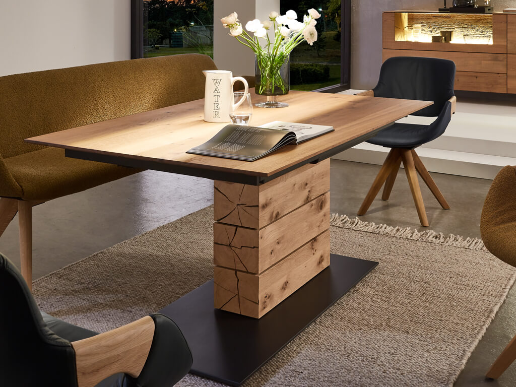 hartmann Naturzeit - Esstisch mit Schweizer Kante & Pöttkerauszug | T100 Esszimmertisch aus Holz mit dekorativer Vase, Buch und Tasse auf dem Tisch, umgeben von gemütlicher Polsterbank und Stühlen