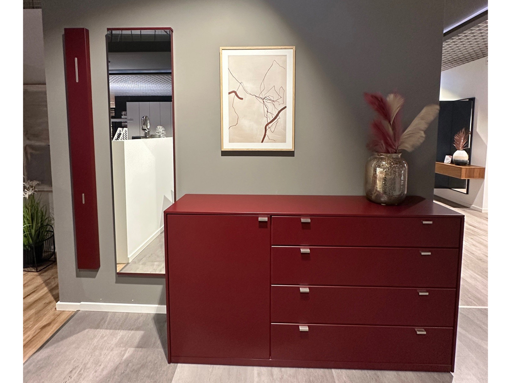 Elegante rote Kommode mit vier Schubladen, flankiert von einem Spiegel, dekoriert mit einem Gemälde und einer Vase, in einem modernen Raum auf grauem Hintergrund platziert.