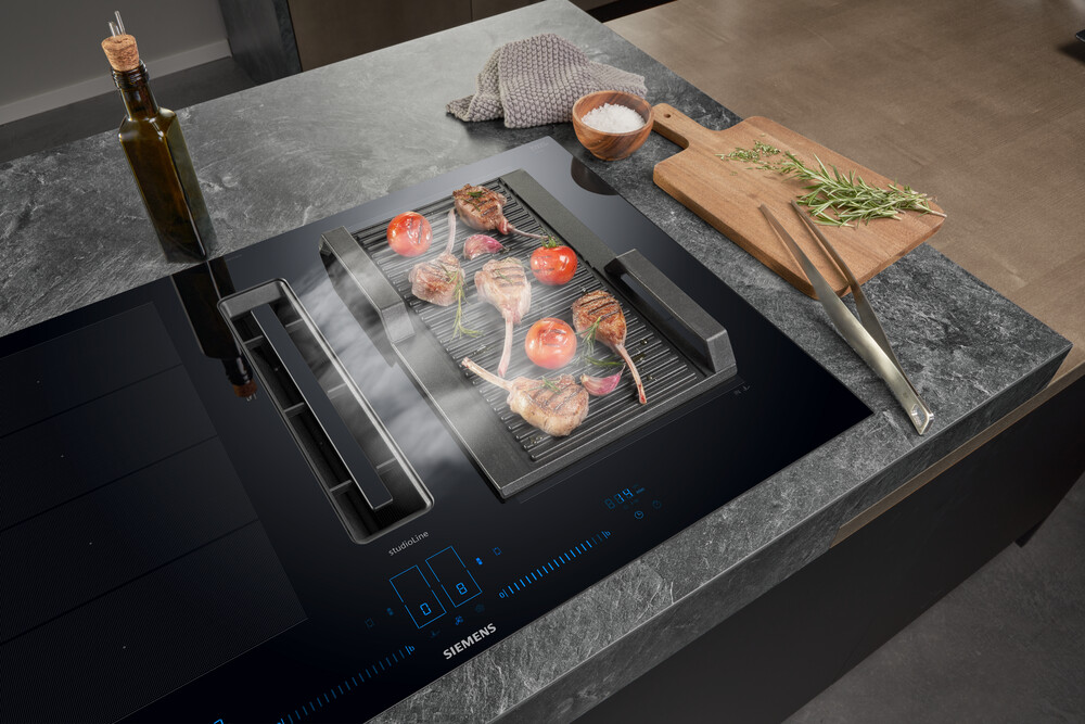 Induktionskochfeld mit gegrilltem Fleisch, Tomaten und Kräutern, umgeben von Kochutensilien