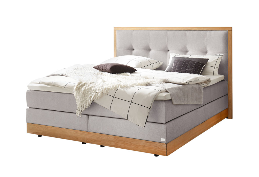 graues Boxspringbett mit braunem Holzrahmen, stilvoll dekoriert mit Decken und Kissen vor weißem Hintergrund