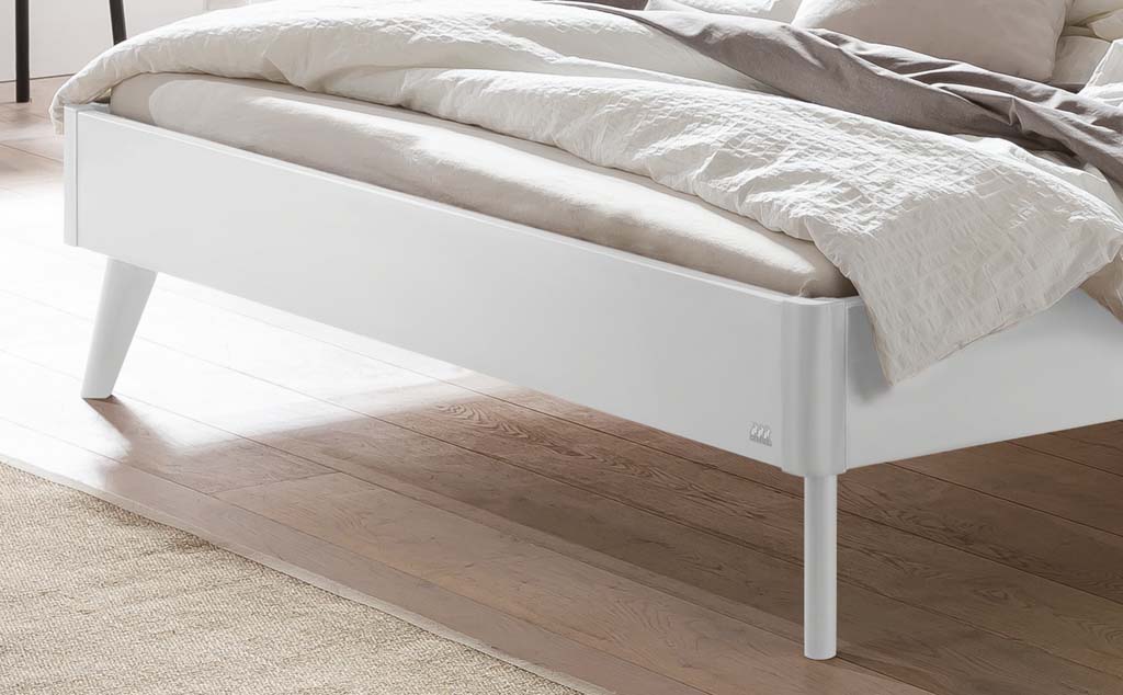 Detailaufnahme eines weißen Holzbettes mit Holzfüßen in einem hellen Raum