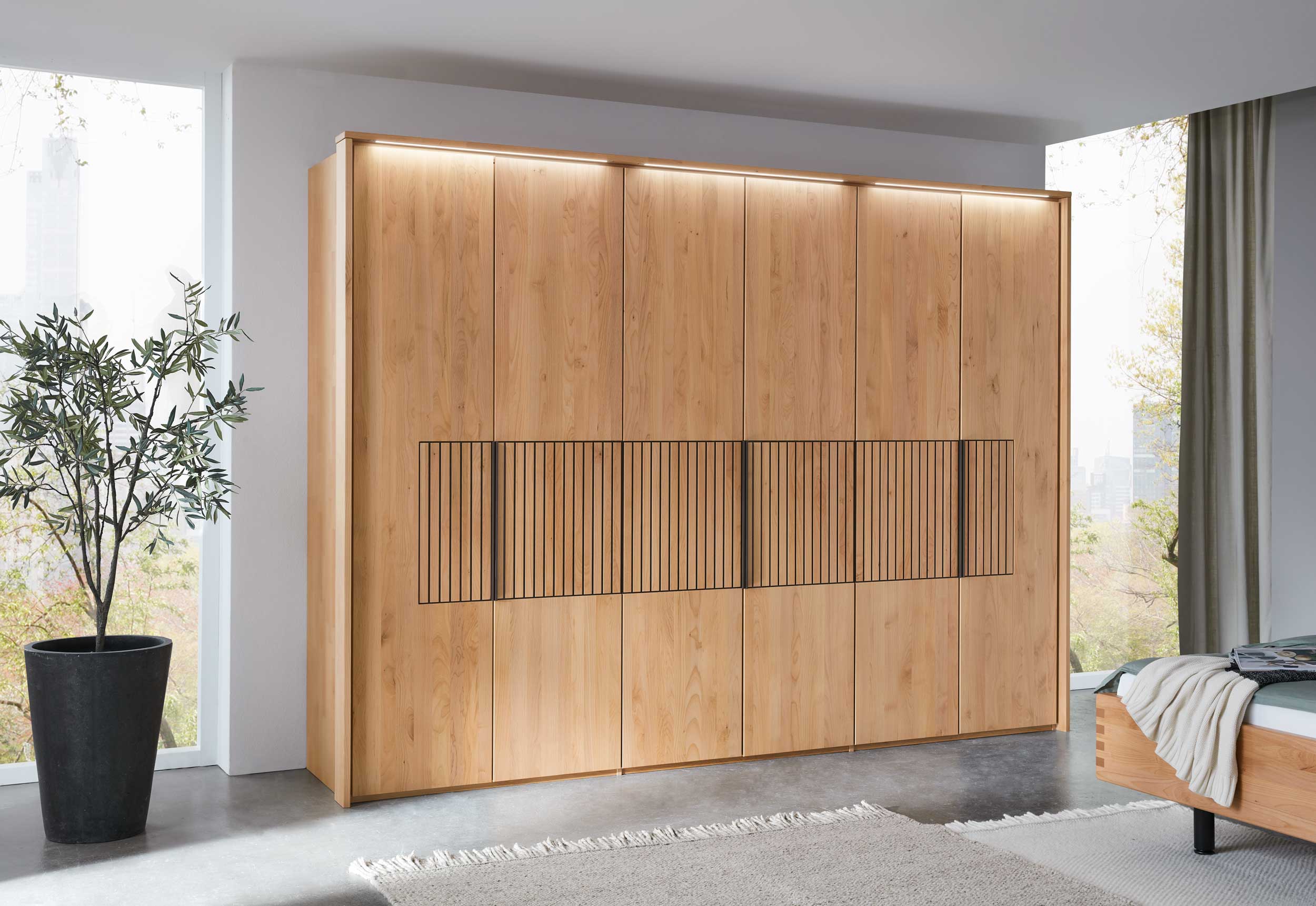 Wöstmann WSM 4400 - Kleiderschrank mit Lamellenakzent | Erle massiv | alle Breiten konfigurierbar Großer, eleganter Holzkleiderschrank mit Beleuchtung in einem modernen Schlafzimmer neben einem Fenster mit Aussicht auf die Stadt