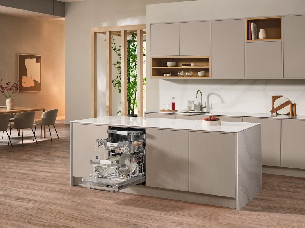 Ein Miele G 7191 SCVi AD Geschirrspüler ist in eine moderne Kücheninsel in beigefarbenen und grauen Tönen integriert, die Tür ist geöffnet und zeigt beladene Geschirrkörbe. Im Hintergrund ist ein Essbereich und ein großer grüner Pflanze zu sehen.