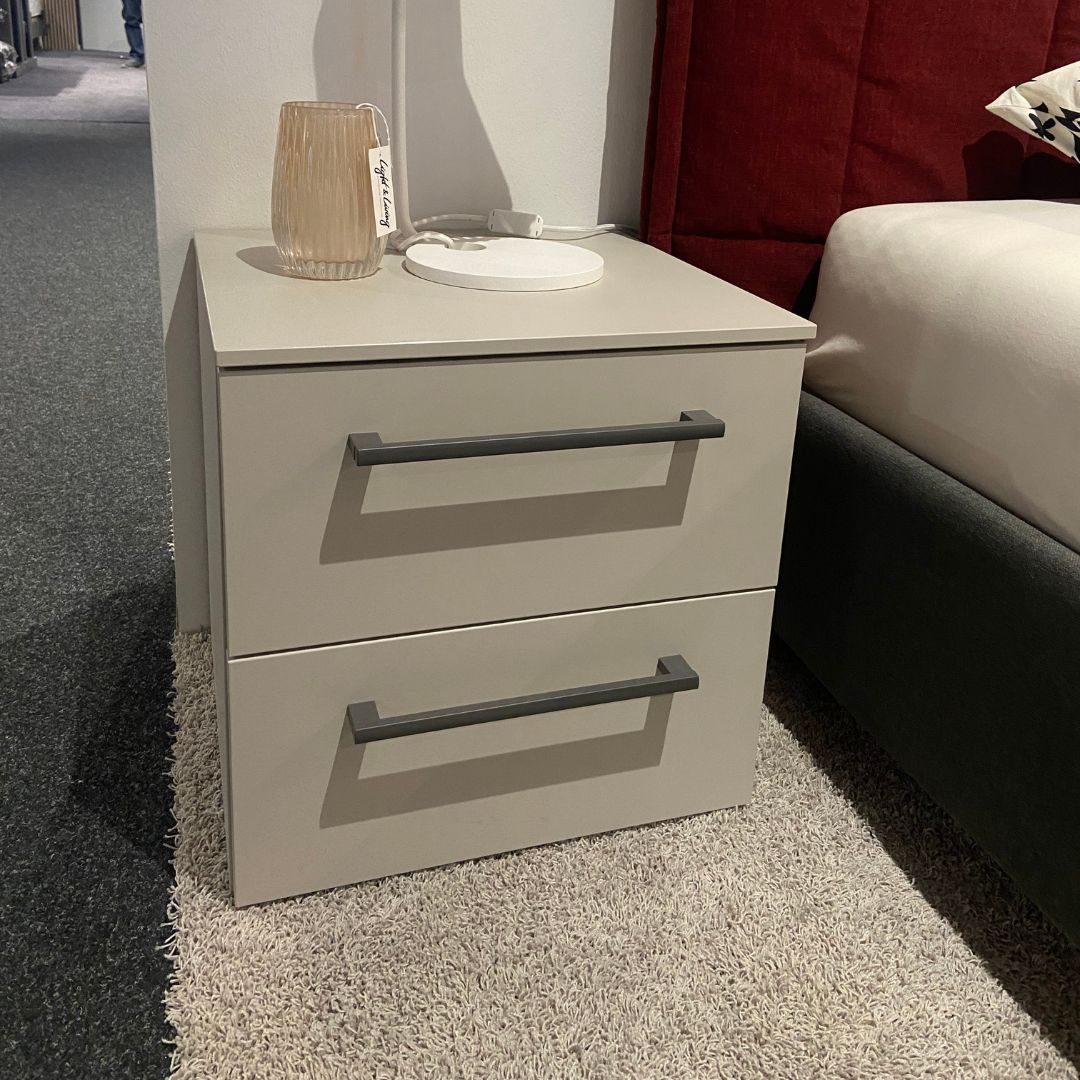 Loddenkemper - MAXX | Nachtkonsole Grauer Nachttisch mit zwei Schubladen und einer Tischlampe darauf, neben einem rot bezogenen Bett, auf einem Teppichboden