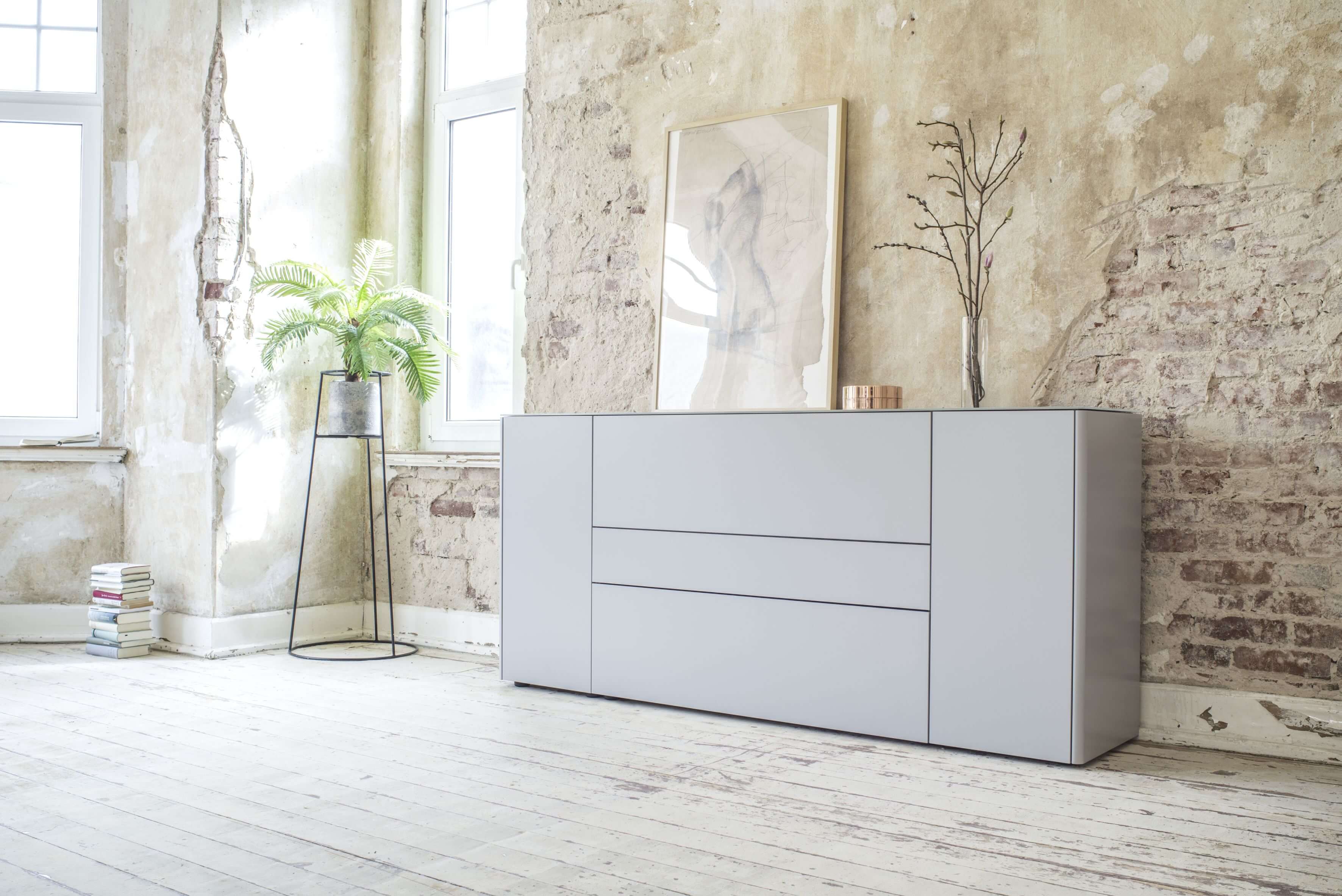 Sudbrock Goya - Sideboard | 2 Türen, 2 Schubladen, 1 Klappe | Glattlack canagrau | B: 205,2 cm Eine schlichte, graue Kommode steht an einer Wand im industriellen Stil mit einem Pflanzenständer und einem Kunstwerk.