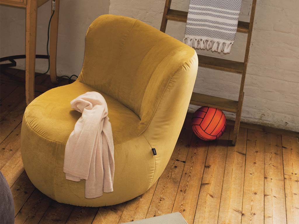 freistil Rolf Benz 173 - Sessel | alle Farben konfigurierbar Ein gelber Sessel auf Holzboden, darauf liegt eine beige Decke. Im Hintergrund ist eine Holzleiter mit einem Handtuch.