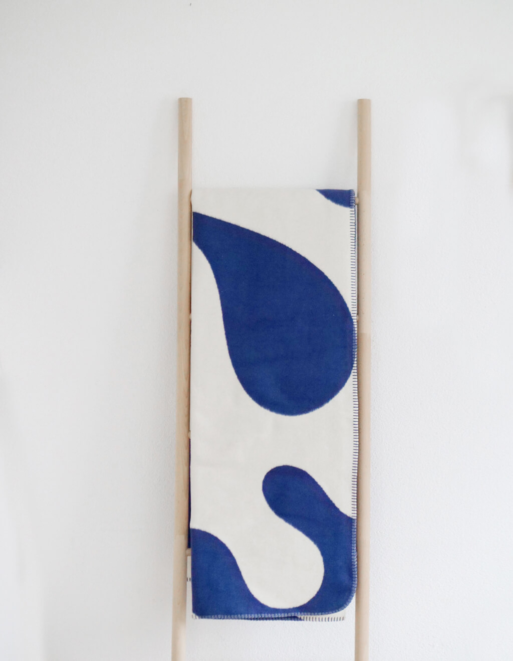 RIEMA LINA – Wohndecke, Bio-Baumwolle - große Muster | 150 x 200 cm | blau-natur Eine weiche Decke in Blau-Weiß mit abstraktem Muster, drapiert auf einer schmalen Holzleiter gegen eine weiße Wand.