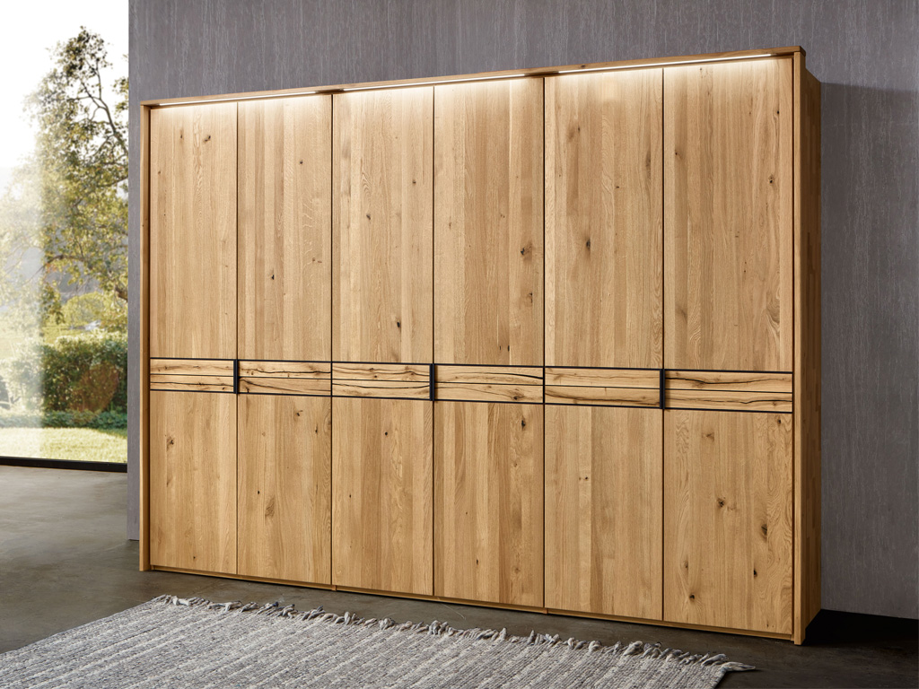 Wöstmann WSM 4300 - Kleiderschrank mit Akzent | Wildeiche massiv | alle Breiten konfigurierbar Großer Kleiderschrank aus hellem Eichenholz mit integrierter Beleuchtung in einem modernen Raum