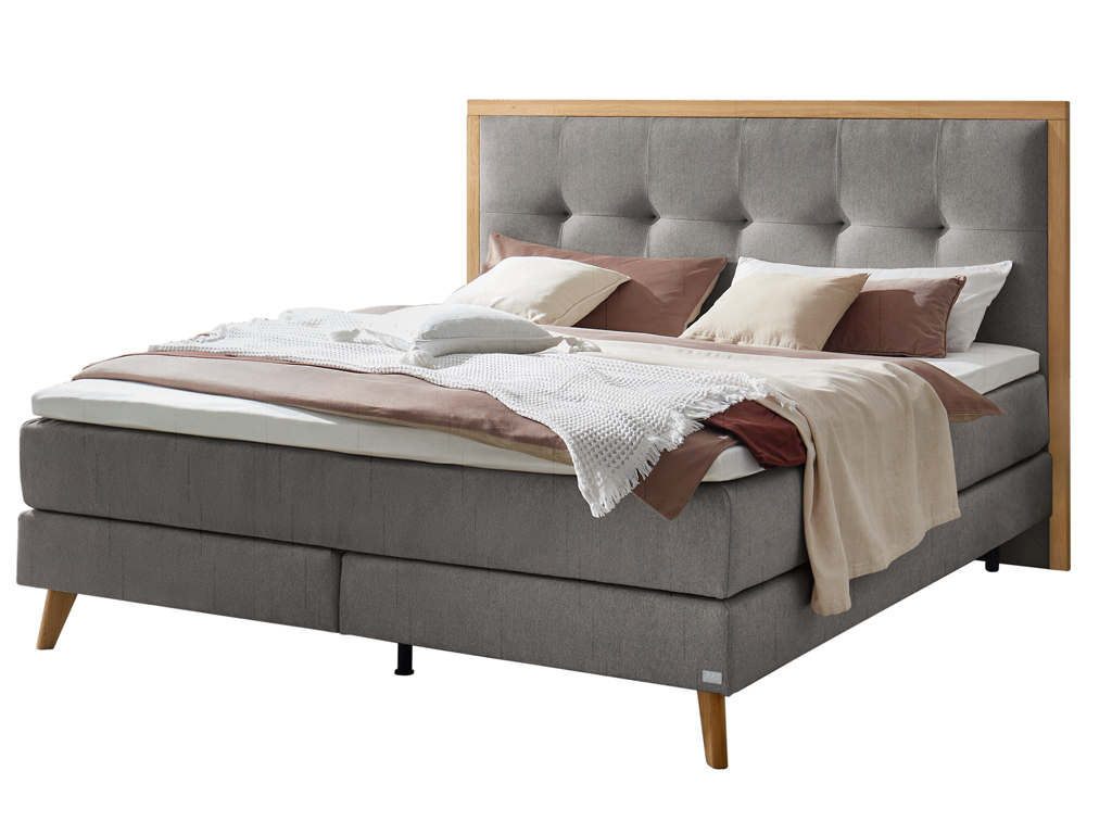 Ein Boxspringbett in dunklem Grau mit einem hellen Holzrahmen um das gepolsterte Kopfteil, freigestellt auf weißem Hintergrund.