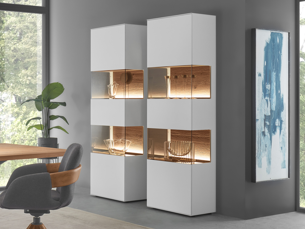 Zwei moderne, hohe weiße Vitrinen mit Holzrückwänden und Beleuchtung, die in einem Esszimmer oder Wohnbereich stehen. Im Vordergrund ist ein Teil eines Tisches und eines Stuhls zu sehen, während im Hintergrund ein Fenster mit Blick ins Grüne. 