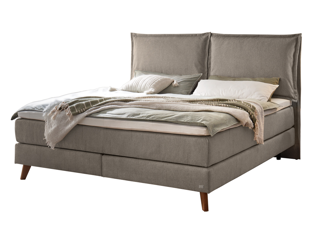 Ein Hasena Boxspringbett Pronto Ricarda mit dunkelgrauem Stoffbezug, das mit Kissen und einer Decke dekoriert ist.