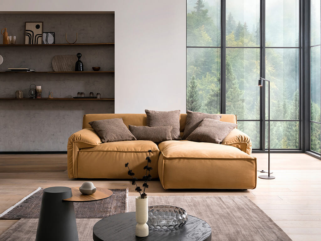 Ein modernes Wohnzimmer mit einem senfgelben Ecksofa, dekoriert mit braunen Zierkissen, vor großen Fenstern mit Blick auf einen Wald. Im Hintergrund ist eine Nische mit Holzregalen und Dekorationen zu sehen.