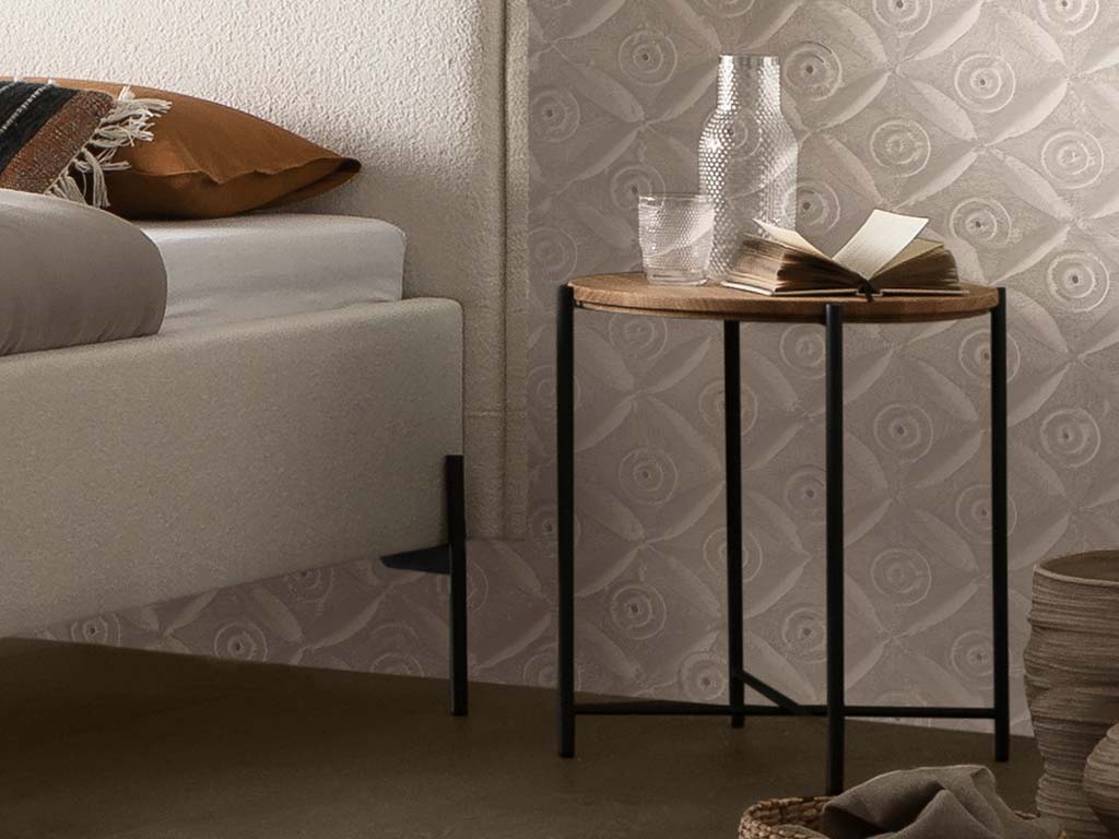 Kleiner brauner beistelltisch mit runder platte auf einem vierfüßigen metallgestell in schwarz stilvoll dekoriert neber einem bett in einem hellen zimmer nali von hasena