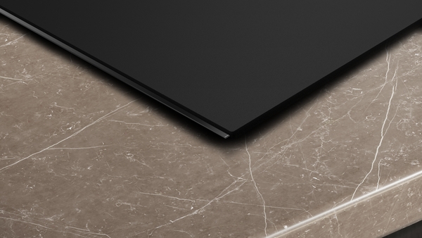 Detailaufnahme der schmalen Kante eines schwarzen Induktionskochfelds, das nahtlos und bündig in eine braun-marmorierte Arbeitsplatte eingelassen ist.