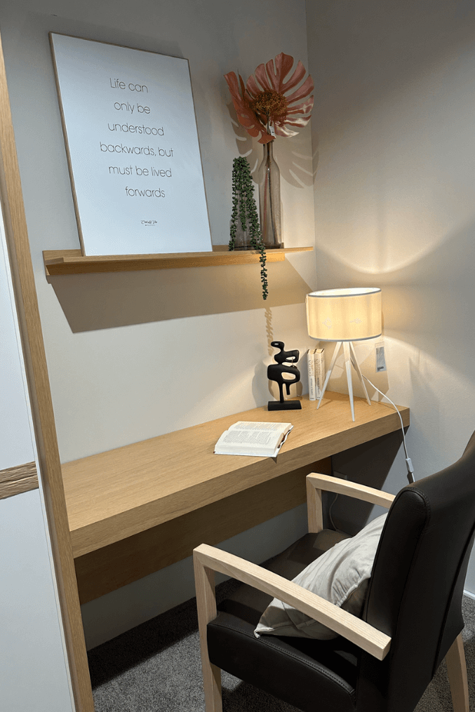 Schreibtischecke mit Holzplatte, Tischleuchte, offenem Buch, Kunstobjekt und Leinwandbild mit Motivationszitat auf Regal, dekoriert mit Blume und Pflanze im Topf.