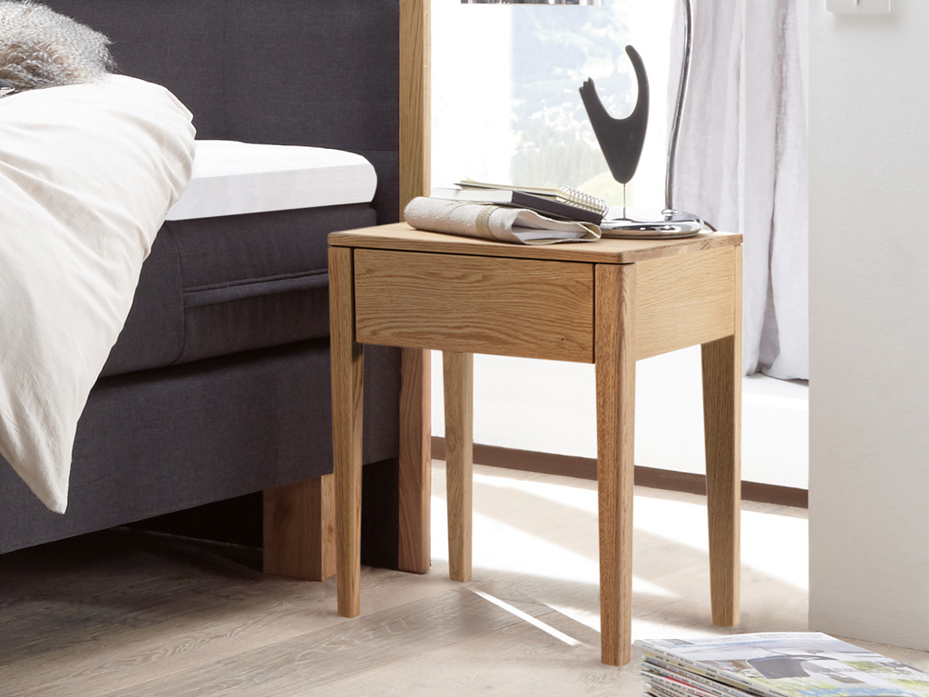 Nachttisch mit vier hohen Beinen und einer Schublade aus hellbraunem Holz vor einem grauen Polsterbett stilvoll dekoriert Chilly von Hasena