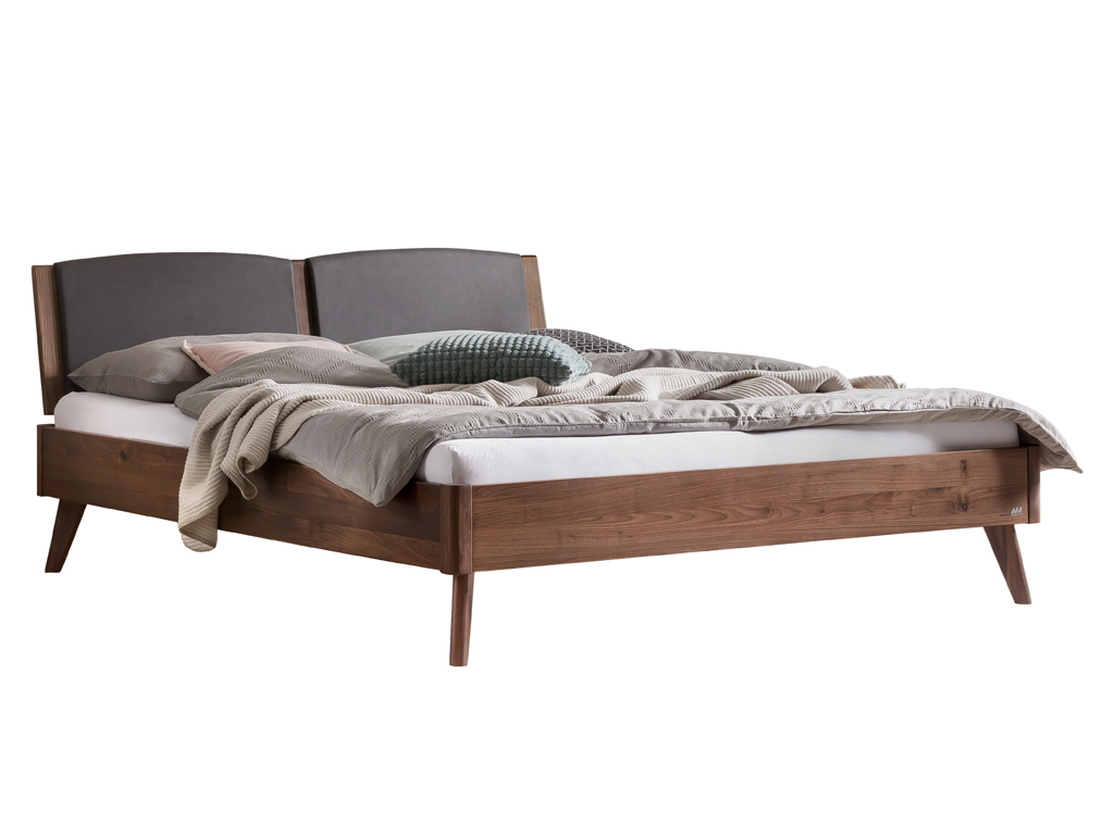 Ein Ansichtsfoto des Hasena Mareto Bettes in der Ausführung Nussbaum (dunkles Holz) mit einem Kopfteil aus Stoff in der Farbe Kul-smoke (dunkelgrau). Das Bett ist mit Bettwäsche, Kissen und einer Decke dekoriert und steht vor einem weißen Hintergrund.