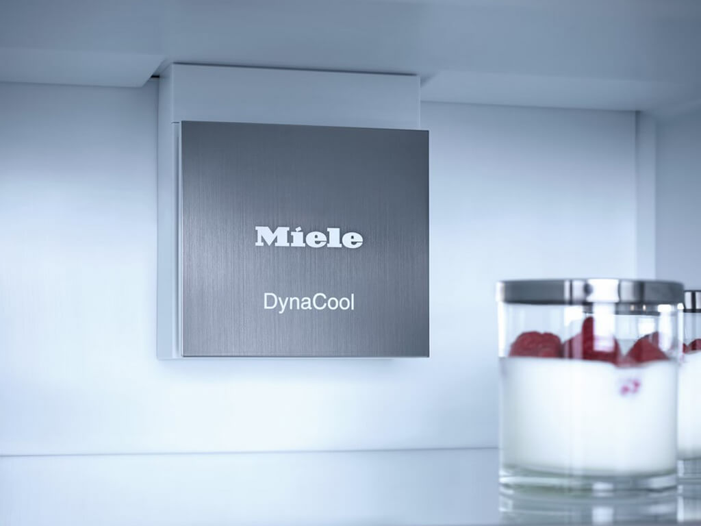 Eine Nahaufnahme des Innenraums eines Miele Kühlschranks, die das silberne „Miele DynaCool“ System an der Rückwand zeigt. Im Vordergrund sind zwei kleine Glasbehälter mit roten Früchten zu sehen.