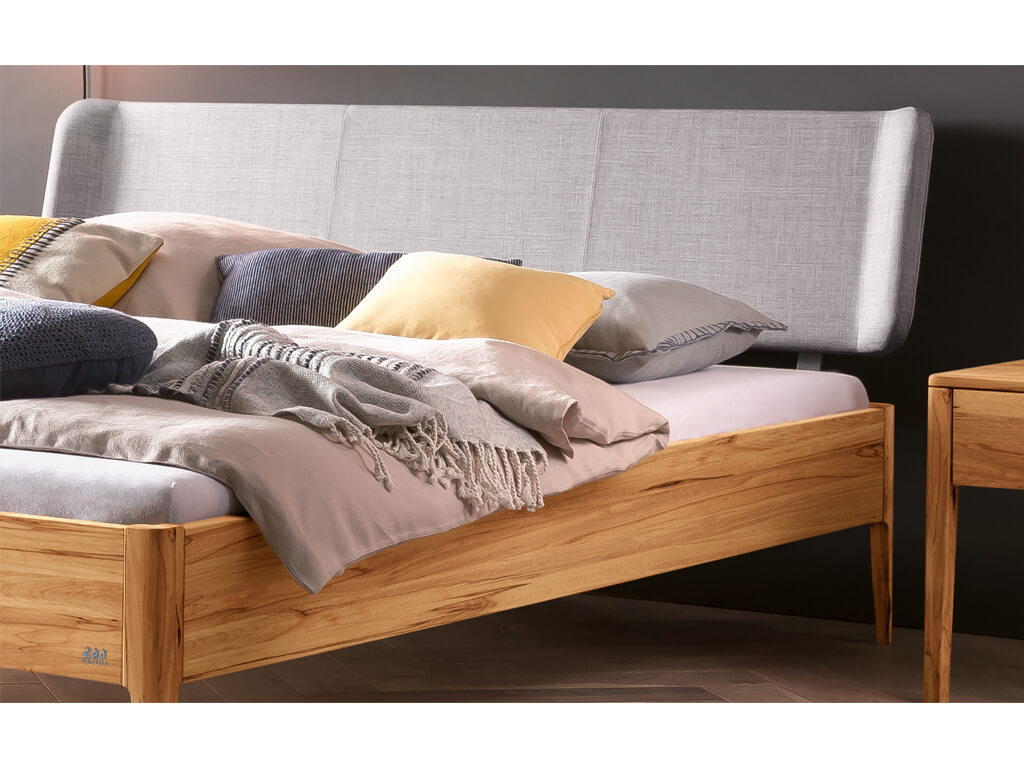 Eine Nahaufnahme des oberen Teils eines Bettes Pienza aus Wildbuche. Das Bett hat ein gepolstertes Kopfteil in hellem 'Pepe-grey' Stoff und ist mit verschiedenen Kissen in Beige, Grau und Gelb sowie einer gestrickten Decke dekoriert.