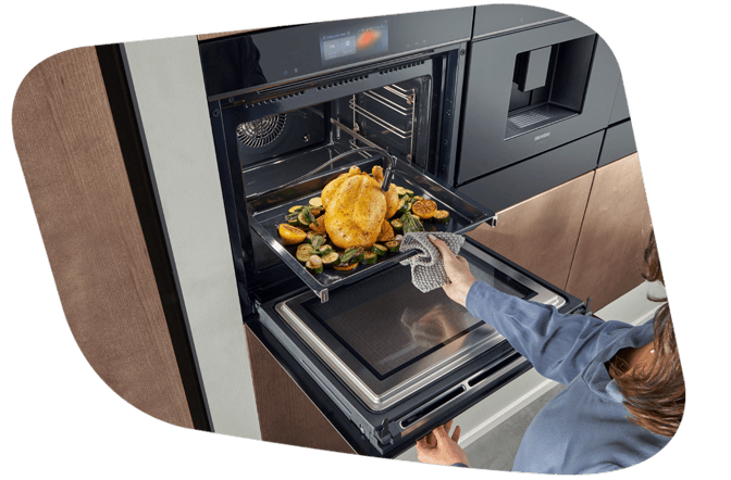 Eine Person entnimmt mit einem Küchentuch einen Braten mit Gemüse aus einem geöffneten Siemens Dampfbackofen.