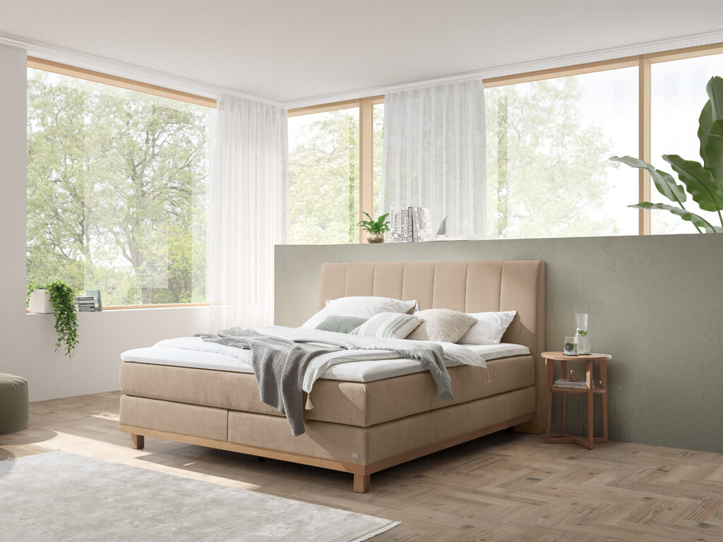 Das Boxspringbett in einem Schlafzimmer mit großen Fenstern, einem Holzboden und einer natürlichen Einrichtung.