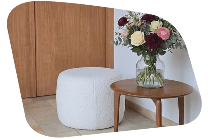 Freistil Pouf 173 in Teddy Creme & i Vordergrund steht ein Holztisch mit einer Vase und bunten Blumen. 