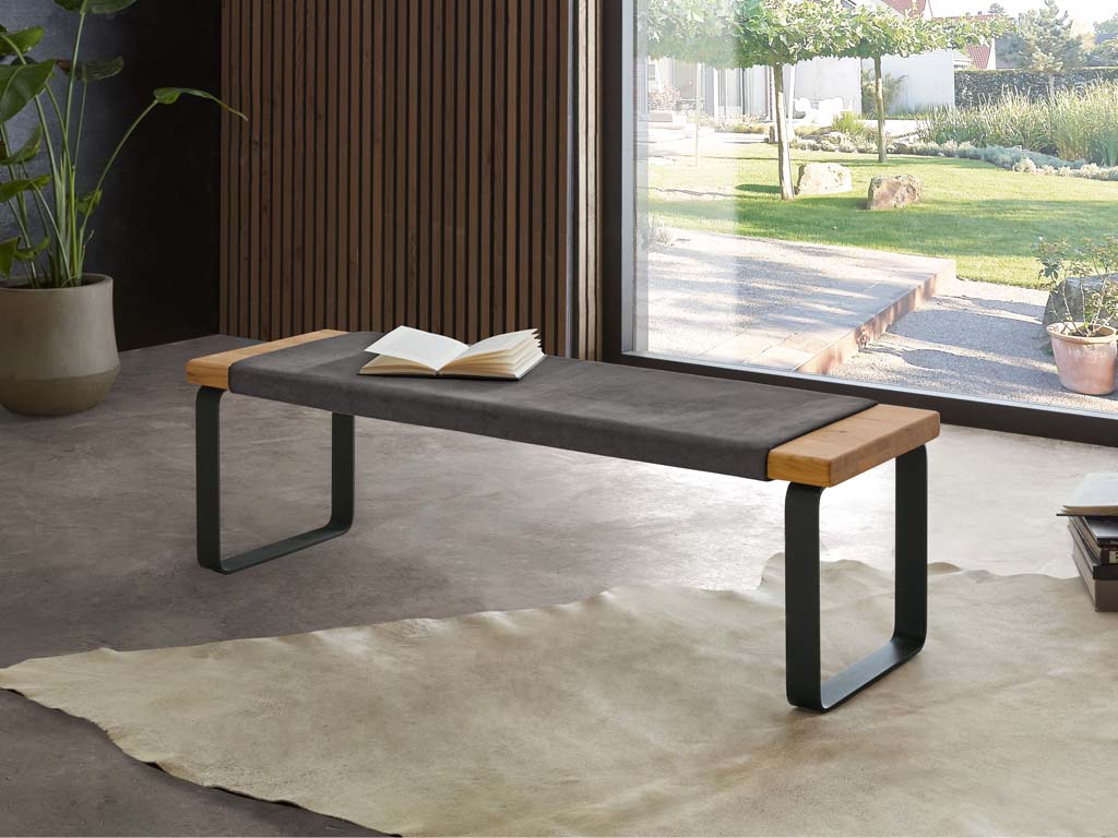 Wöstmann WSL 8800 - Sitzbank (1482) | Wildeiche massiv | B: 120,0 cm Sitzbank mit Holz und Metallrahmen, dekoriert mit einem offenen Buch, vor einer großen Fensterfront mit Blick in den Garten
