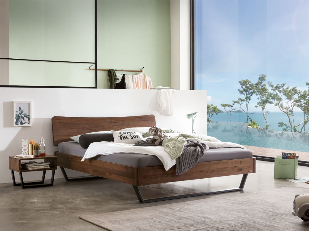 Ein Hasena Bettmodell Tresali aus Nussbaum, das in einem hellen, modern eingerichteten Schlafzimmer mit großen Fenstern steht.