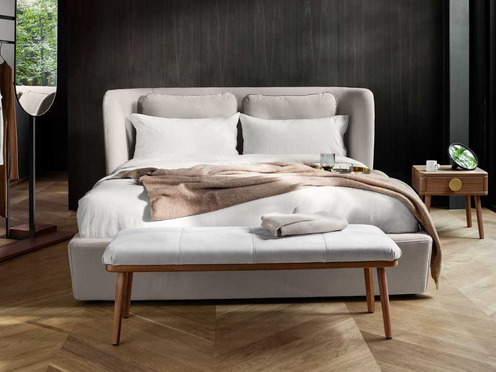 Ein stilvoll eingerichtetes Schlafzimmer mit einem großen Doppelbett, umgeben von dunklen Holzwänden, einem gepolsterten Hocker und einem Nachttisch mit Dekorationen.