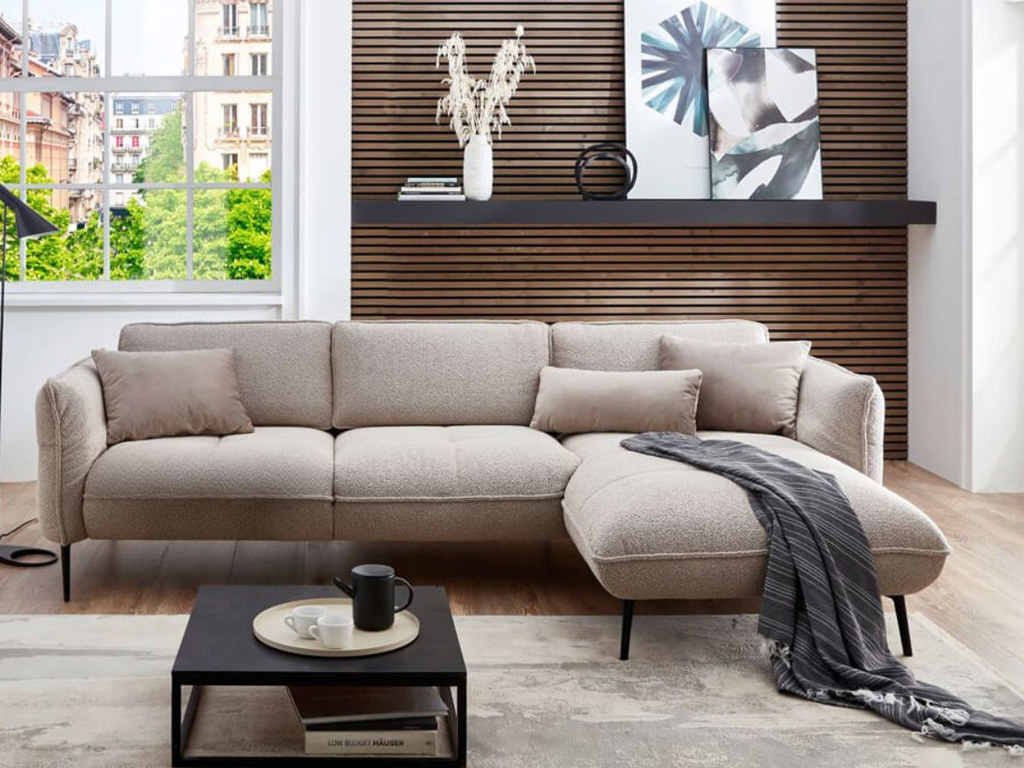 Helles Wohnzimmer mit einem beigefarbenen Ecksofa, dekorativen Kissen und einer Decke, im Hintergrund eine Holzlattenwand mit Pflanzen und Bildern.